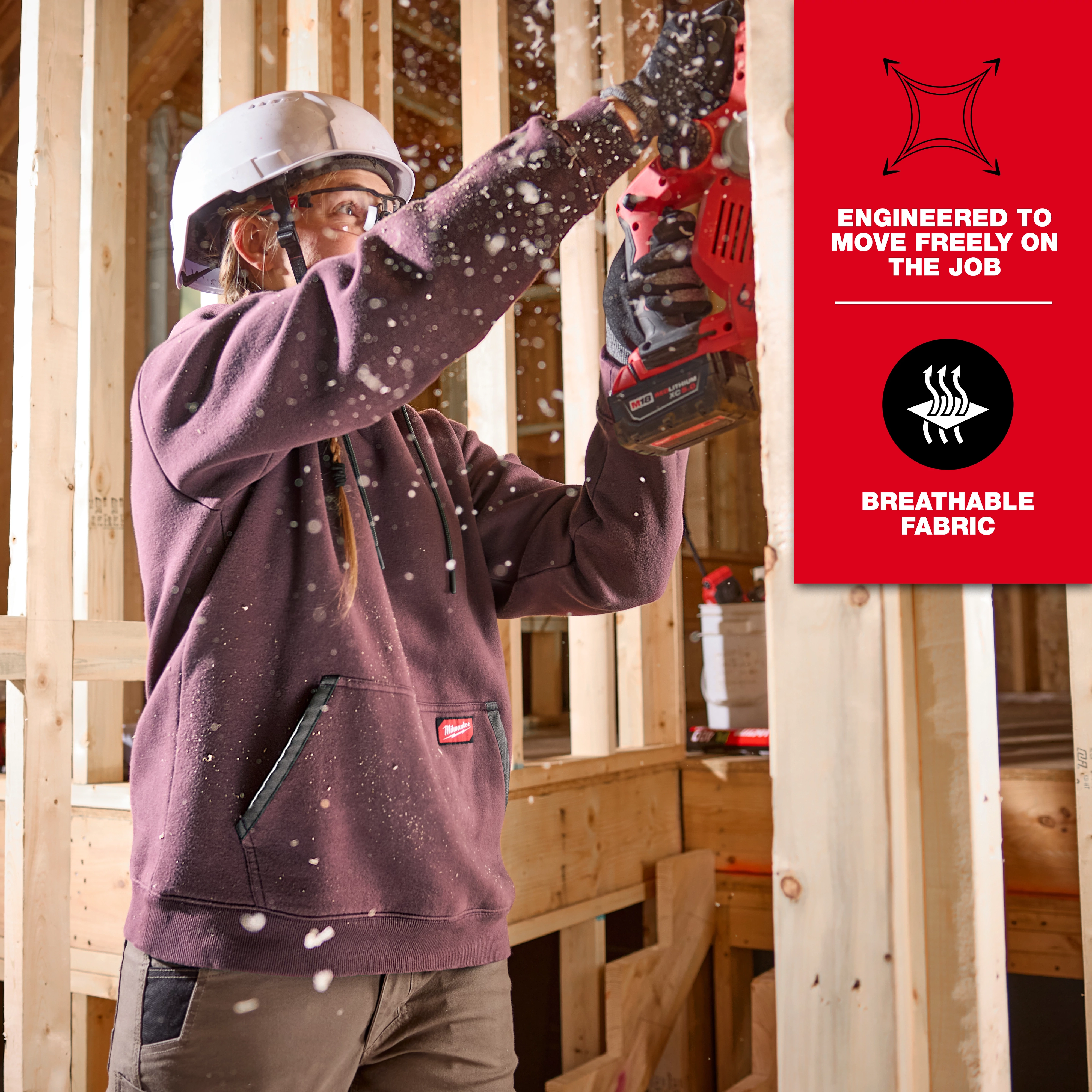 A woman in a hard hat is using a power tool on a construction site. She is wearing a Women's FREEFLEX™ Pullover Hoodie. The hoodie is engineered for free movement and features breathable fabric. There is sawdust in the air, and the background shows wooden framing.