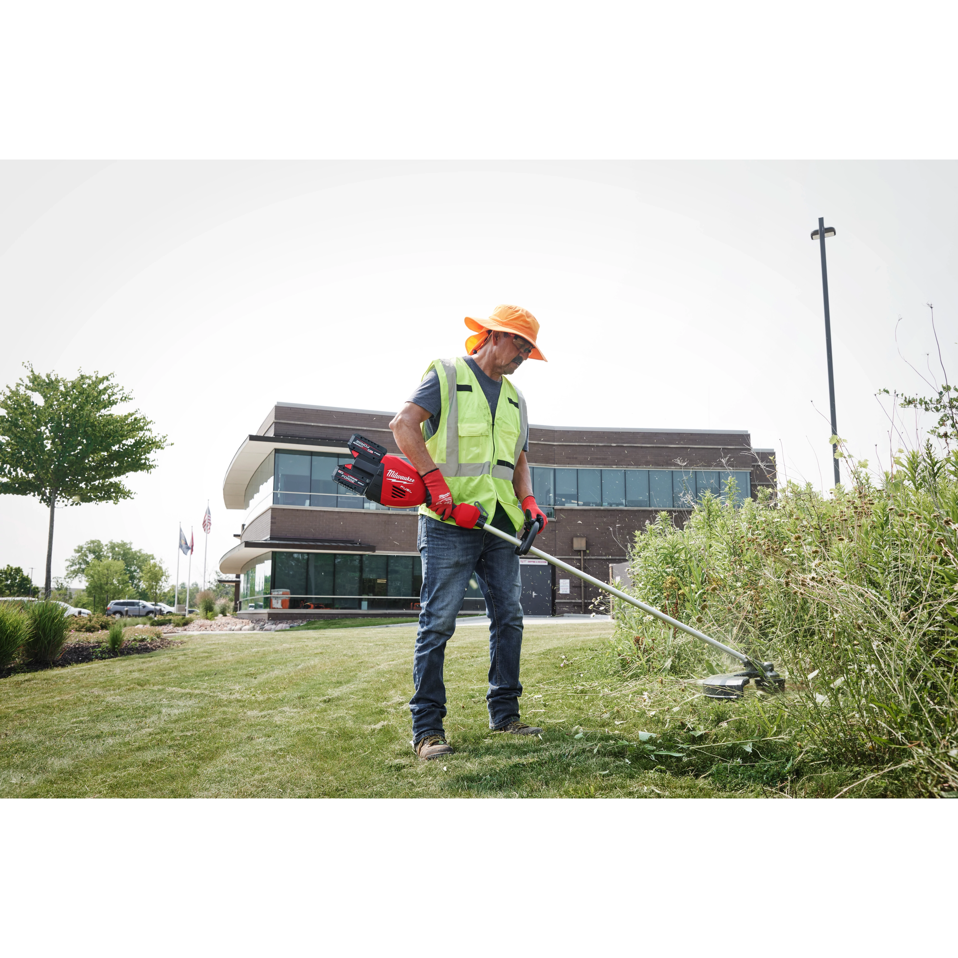 A person in a safety vest and gloves trims grass with a weed cutter outside a modern building.