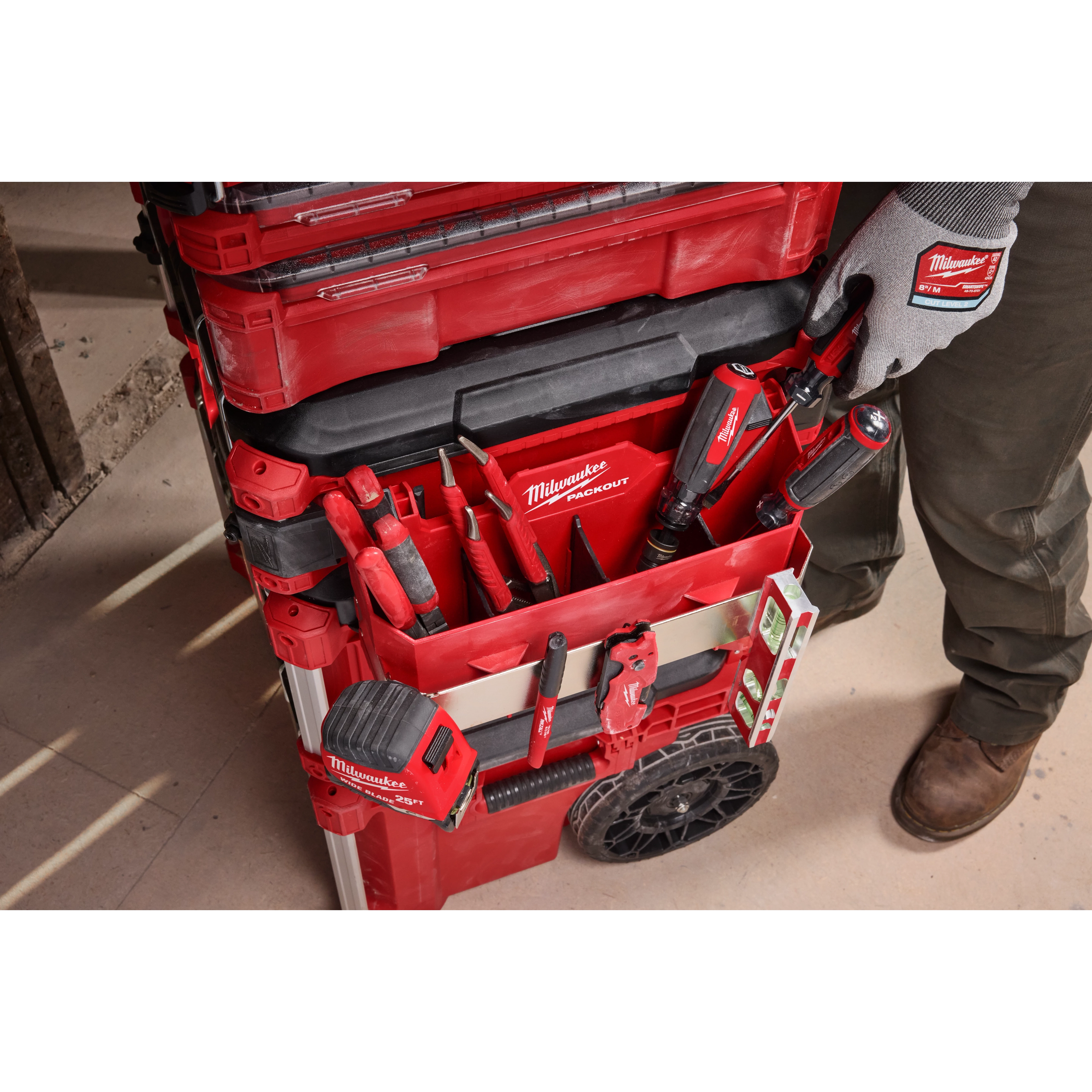 The image shows the PACKOUT™ Tool Box Caddy Attachment connected to a stack of red Milwaukee PACKOUT™ toolboxes. The caddy holds various Milwaukee tools, such as screwdrivers and pliers, and includes organization slots and pockets. A person wearing gloves is reaching for a screwdriver.